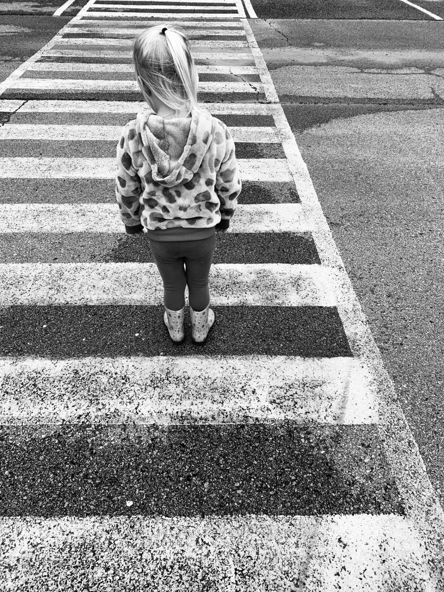 Black-and-white photograph of a young girl standing on a painted crosswalk, seen from behind